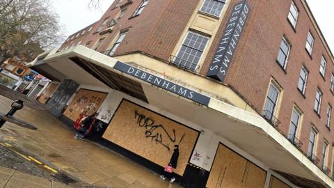 A boarded-up entrance to Debenhams in Norwich. Two people are walking past the graffiti-covered boarding.