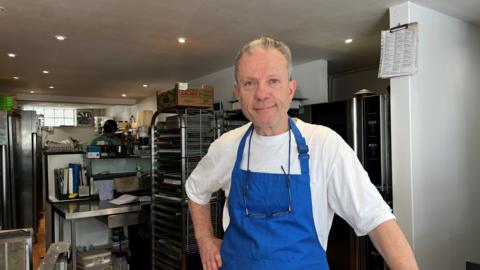A man in a blue apron and white shirt stood in a bakery kitchen.