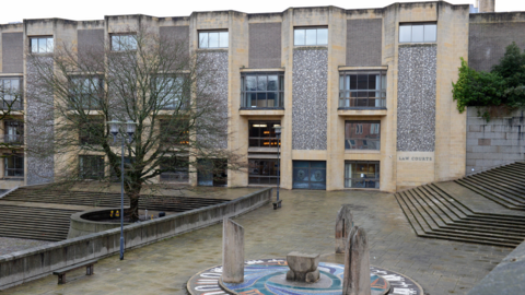 Winchester Crown Court, a plain building resembling an office block, sits beyond a courtyard with a bare tree and an art feature with three small wooden pillars on a mosaic. To the right, a set of long steps ascend until they disappear out of shot.