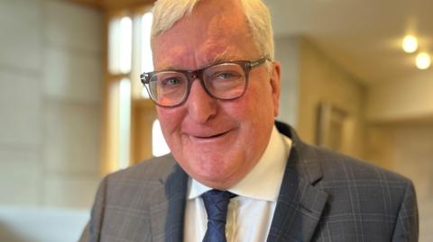 Fergus Ewing has short white hair. He is wearing glasses and a grey suit over a white shirt with a blue tie. Is pictured indoors.