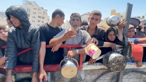 Tayseer Naeem, an older man with a grey beard, holds a saucepan amidst a line of people behind a metal fence at a soup kitchen in Gaza.