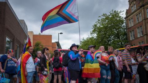 Newcastle Pride 2024. A crowd of people line the street. Many are holding rainbow flags and are dressed in colours associated with Pride.