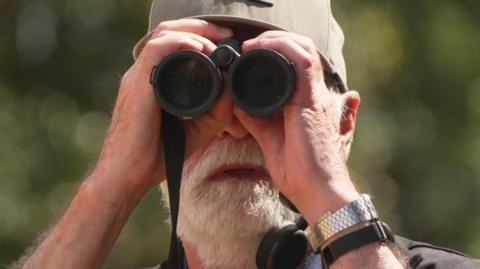 Nigel Hewitt is holding a black pair of binoculars and wearing a cap. He has a white beard.