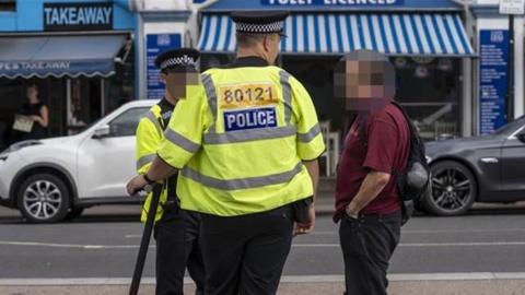 Two police officers wearing their uniform holding an e-scooter bike and a civilian standing next to it but his face has been blurred. Behind them is a main road with parked cars and food shops and stalls.