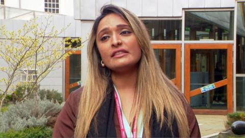 A woman with long fair hair pictured in a garden. She is wearing a red jacket and has gold nose and ear jewellery. 