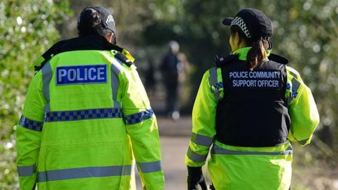 Generic image of two female police officers in uniform wearing high vis jackets, one saying 'police', the other with a police community support officer black vest over the top. They are walking away from the camera down a lane with trees either side