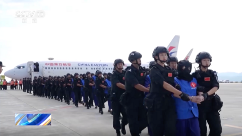 Rows of security personnel in black clothes and helmets escorting suspects from a plane. Each suspect, wearing bright blue attire and a black mask over their heads, is flanked by two guards.