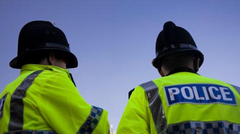 Two police officers stand with the backs to the cameras. They are both wearing high-visibility jackets and black helmets. Both jackets have "POLICE" written on the back. 