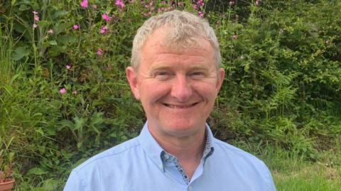 Steve Rubidge is smiling at the camera as he stand in a garden. He has short grey hair and is wearing a pale blue shirt. There are ferns and pink flowers in the hedging behind him.