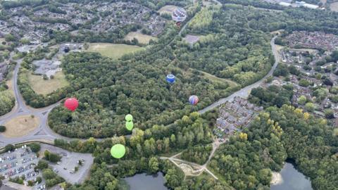 A birds eye view of multi-coloured hot air balloons in the sky while trees, a car park and buildings are below.