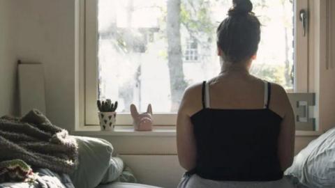 A woman looks out of a window with her back to the camera. She is sitting in a bedroom.