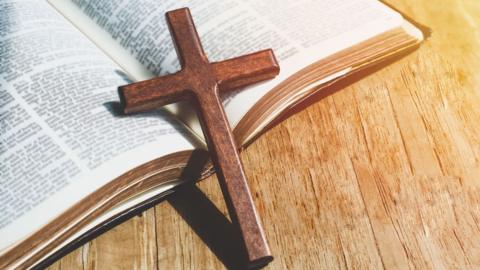 Closeup of a wooden Christian cross necklace on a Bible. There is subtle sunlight coming from the right of the picture. 