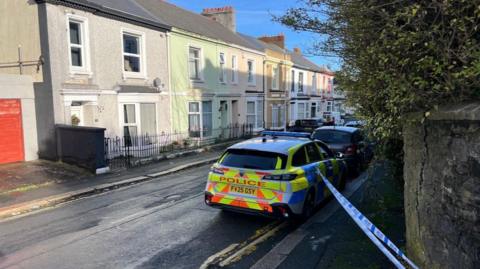 A picture of a police cordon on near Mutley. There is blue police tape and a police car parked behind it. 