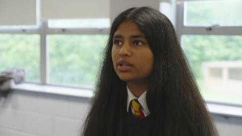 A girl in a school uniform with a yellow, red and blue tie and black sweater sat in a classroom