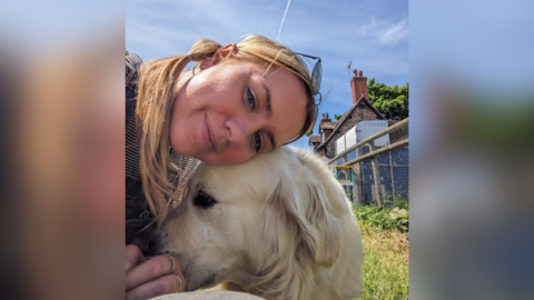 Rhianne Aitken smiling with a golden retriever.  A fence and a house are in the background. The image was taken on a sunny day.