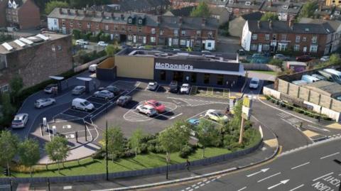 A single storey rectangular building with a white McDonald's sign on its roof stands in the centre of the plot off a main road. the plot has a number of car park spaces and some landscaping close to the road.