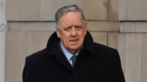 William Lloyd-Lavery, a man with grey hair, a dark jacket, blue shirt and blue tie, pictured beside a grey brick building. He has his mouth slightly ajar.