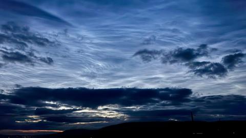 A denim blue sky is lit up by silvery wisps of cloud. The sun is breaking through below the lower clouds and casting a faint golden light.