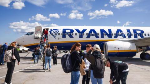 Against a blue cloudy sky, a group of travellers descend a Ryanair plane