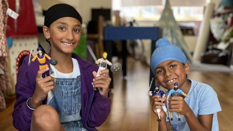 A girl and a boy holding an action figure in each hand, smiling at the camera in the room of a house