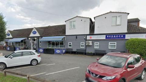 A white car and a red car parked in a car park in front of a building. The building is mainly single storey with some dormer rooms. There are blue and white striped awnings over outdoor seating areas on the left side of the building. On the right side there are benches against the wall of the building. A blue sign on the front of the building says Redbeck Hotel.