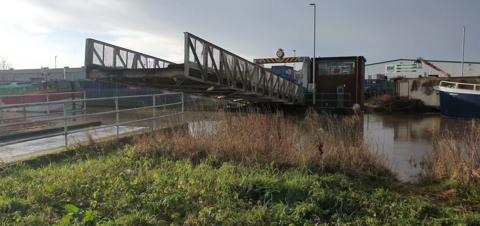 A metal lift bridge in operation. There is a control room and a height barrier at the far end of the bridge. The end nearest to the camera is lifted above its normal position.