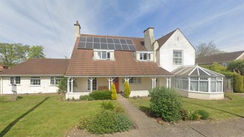 The exterior of a care home. It is a mixture of a single-storey and two-storey building with a glass conservatory on the right. It has white walls and a tiled pitched roof with solar panels on it.
