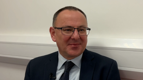 A photo of Mike Cutland in a suit in an office environment. He is smiling. He has glasses and short brown buzzed hair. 