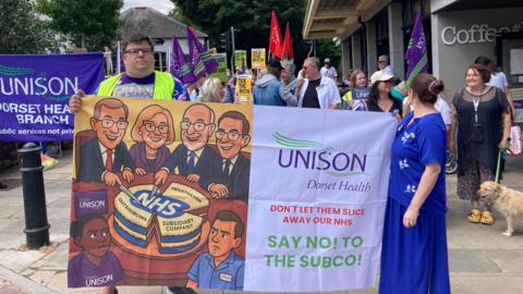 A group of people holding unison flags in a town square. A man in a high visibility vest and a woman in a blue dress stand at the front, holding a large banner with the Unison Dorset Health logo and reading "don't let them slice away our NHS, say no to the SubCo"