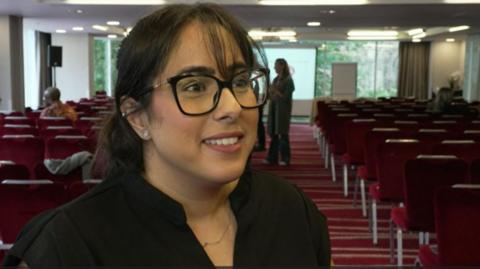 Amarjit standing in the foreground of a large conference or meeting room. She is wearing a black top, dark-framed glasses, small stud earrings, and a thin necklace. Her hair is tied back, with a few strands falling across the forehead. The room contains many rows of red upholstered chairs arranged in neat lines facing the front. A red patterned carpet with horizontal stripes runs through the room. The lighting is bright, with multiple ceiling lights illuminating the space.