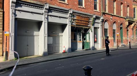 A police officer is standing on a road within a cordon outside a two-storey building. Police tape can be seen at one end of the cordon. Wooden doors to the building are open.