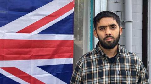 Ibrahim Syed, who has short dark hair and a beard and is wearing a black and grey check shirt, stands outside the Deen centre in Birkenhead, with the union jack he had found tied to railings outside now on display in the window behind him. 