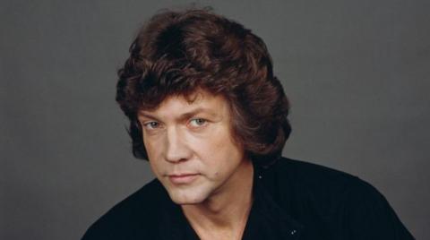 A man with curly brown hair is looking at the camera. He is wearing a dark top.