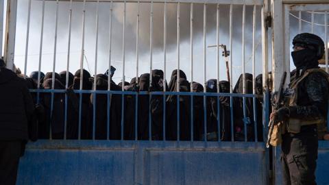 Syrian security force member pictured holding a gun and standing next to a blue metal gate at al hol camp with women in niqabs crowding at the gate
