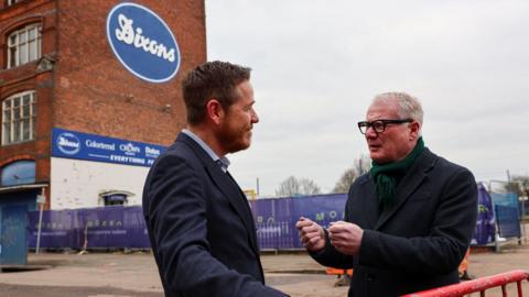 Two men are standing in front of a large red brick building that has a blue sign that reads Dixons. The man on the left has short brown hair and is wearing a dark blue suit jacket with a light blue shirt. The other man has short white hair and black glasses, he is wearing a dark jacket and a dark green scarf.