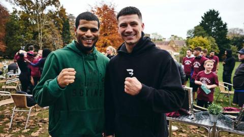 A man in a green hoodie stands next to a man in a black hoodie smiling at the camera in the middle of a park with people in maroon t-shirts in the background. Both have their fists raised in a mock boxing pose