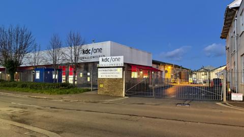The outside of the Swallowfield site, which is a low square building behind a large fence. The KDC/One logo is visible on the building and on the gate. 