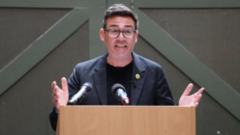 Andy Burnham mid-speech, wearing glasses, suit and tie, with arms stretched out and open palms.