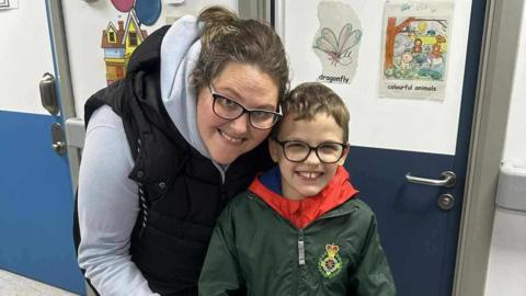 A woman with brown hair and glasses in a white and blue hooded top next to a child with brown hair and glasses in a green top with a yellow badge