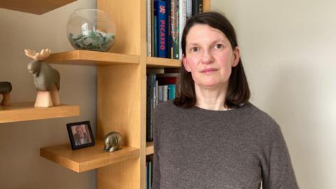 Helen stands in front of a bookshelf, wearing a brown sweater. She has brown, shoulder-length hair and brown eyes. Various figurines and pictures adorn her wooden bookshelf as well as several art books.