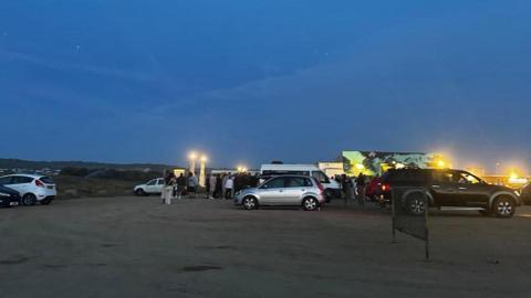 The car park at night. There are cars and a white Ford Transit van parked. Beyond that is a large group of about 50 people all facing one way, with their backs to the camera.