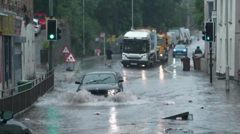 A car driving through flood water, half submerged. There is some rubbish floating in th water.