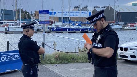 Two police officers stood by Ipswich Waterfront with boats and warehouses in the background