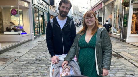 Clinton, Bee and Mabel stand in the highstreet in St Peter Port. Clinton is wearing a black zipped jumper with a white t-shirt underneath. He has short dark brown hair. Bee is wearing a dark green dress and a light green jacket which is unzipped. She has shoulder length red hair. Mabel is in her pushchair and has a brown ribbon headband on her head.