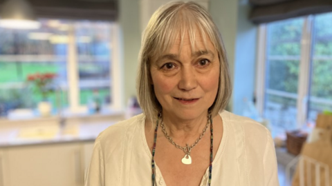 Penny Parry is standing in her kitchen wearing a white shirt. She is smiling.