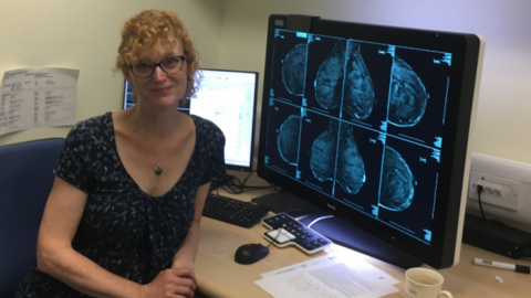 Dr Vinnicombe smiles at a desk, next to a large screen with eight different projections of breast screenings on it. She wears a dark top, and has very curly red hair. 