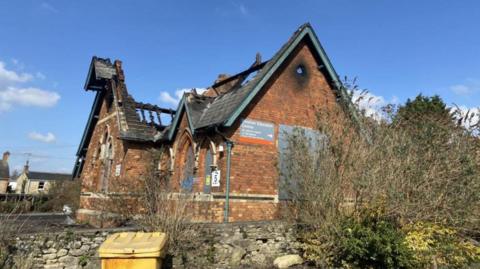 An old school building, missing most of its roof. The red brick around the building is visibly charred, and beams have been exposed by the fire damage. 