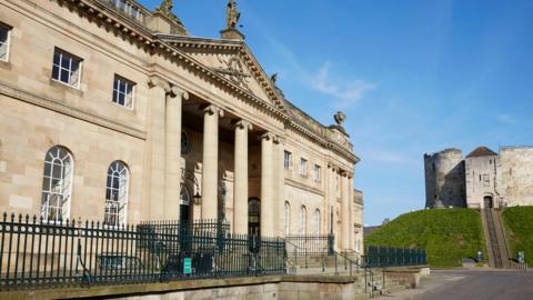 York Crown Court is on the left of the image. It is a large Georgian building of pale stone with iron railings and steps in front. To the right is the mound and Cliffords Tower.