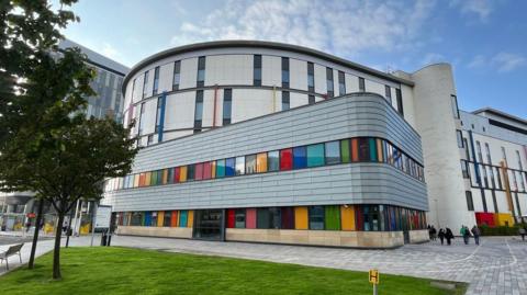 A view of the full building of the Royal Hospital for Children in Glasgow on a clear day. It is a modern building with colourful windows, and grass and trees outside.