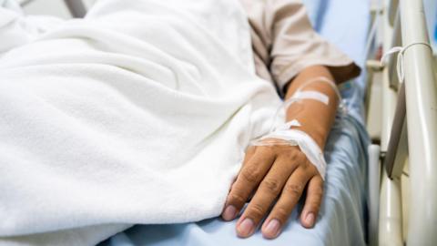 A man lies in a hospital bed with tubes inserted in his arm.